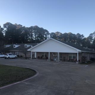 A building with a front porch and a parking area surrounded by trees under a clear sky.