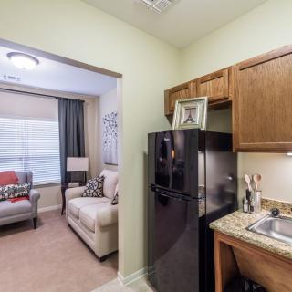 A cozy apartment featuring a compact kitchen with wooden cabinets and a black fridge, opening into a comfortable living room with a couch and armchair.