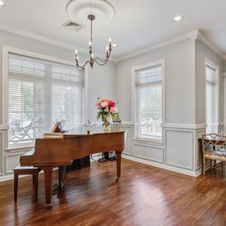 A well-lit living room featuring a grand piano, hardwood floors, and large windows adorned with a floral centerpiece.