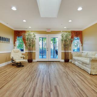 A spacious living room with wooden floors, a comfortable sofa, chair, and potted plants near large windows.