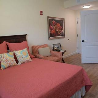 A cozy bedroom with a coral-colored bedspread and an armchair, featuring decorative pillows and a painting on the wall.