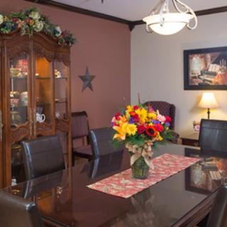A cozy dining room with a table set, decorated with flowers and surrounded by dark leather chairs. A china cabinet is in the corner, and a lamp provides warm lighting.