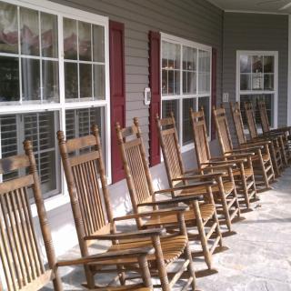 A row of wooden rocking chairs lined up on a porch, facing large windows with red shutters.