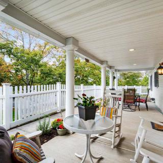A cozy porch area with rocking chairs, a table, and plants, all under a covered outdoor space with a white picket fence surrounding it.