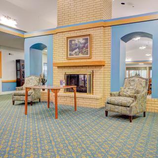 A cozy living room interior with a fireplace, two patterned chairs, and a wooden table on a carpeted floor.