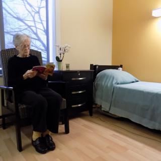 An elderly woman enjoys reading in a cozy room with a bed nearby.