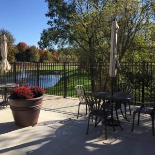 A serene patio setting with tables and chairs overlooking a pond and surrounded by trees on a sunny day.