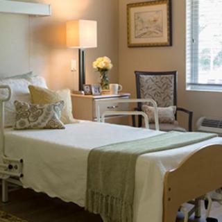 A well-lit hospital room featuring a neatly made patient bed, bedside table with a lamp and flowers, and a comfortable chair by the window.