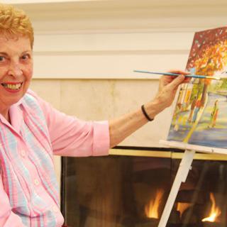 A woman smiling while painting on a canvas at an easel indoors.