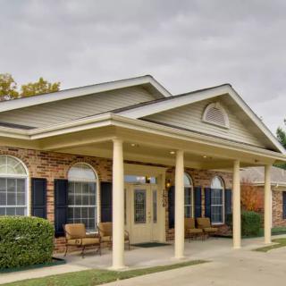 A building with a porch supported by columns and brick walls, featuring several windows and chairs.