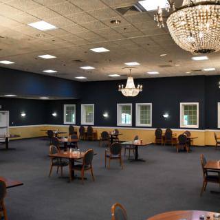 An elegantly lit banquet hall with chandeliers, featuring several tables and chairs placed on a carpeted floor.