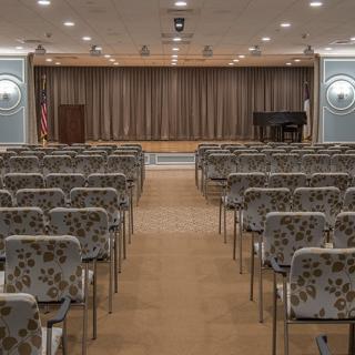 An empty auditorium with rows of chairs facing a stage with closed curtains and a piano.