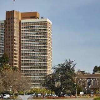 A tall, modern building surrounded by greenery under a clear sky.