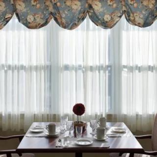 A neatly set dining table with a floral valance and sheer curtains in the background.