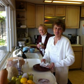 Two women are standing in a kitchen near the sink, engaged in washing dishes. The room is well-lit by daylight coming through a large window.