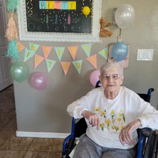 An elderly woman sits in a wheelchair surrounded by colorful decorations and balloons, celebrating a special occasion.