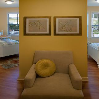 A cozy armchair with a decorative cushion sits against a yellow wall, adorned with framed artwork.