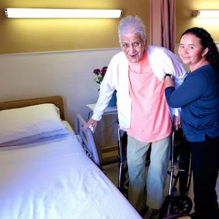 A caregiver assists an elderly person in a hospital room, providing support for mobility.