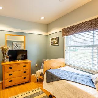 A cozy bedroom featuring a bed with a blue throw, a wooden dresser with a TV, and large windows with blinds.