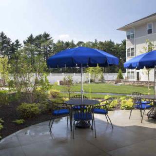 An outdoor patio area with blue umbrellas and metal furniture, set against a landscaped garden.