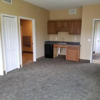 An empty room with a kitchenette, featuring cabinets and a sink, carpet flooring, and open doors.