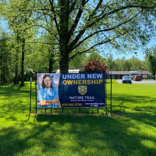 A banner announcing new ownership in a green outdoor setting.