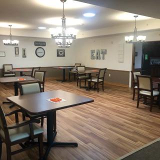 A well-lit dining area with neatly arranged tables and chairs, featuring chandeliers and wooden flooring.