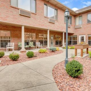 A courtyard area with a brick building, featuring a pathway, benches, and landscaped shrubs.