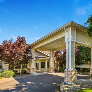 The image shows a building entrance with a covered driveway, landscaped with trees.