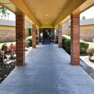 A covered walkway leading to the entrance of a building, flanked by brick columns and landscaping.