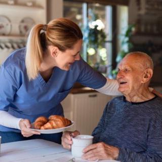 A caregiver serves breakfast to an elderly man, sharing a warm smile in a cozy kitchen setting.