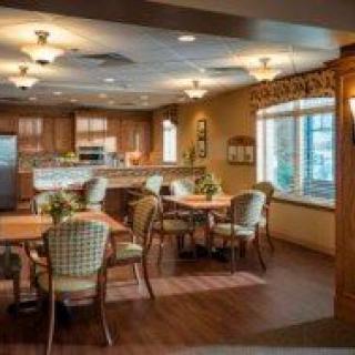 A cozy dining and kitchen area with wooden tables and chairs, warm lighting, and large windows.