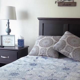 A cozy bedroom setting with a neatly made bed, decorative pillows, and a nightstand featuring a lamp and framed art.