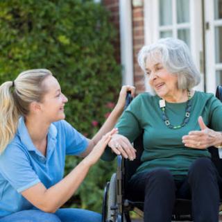 A caregiver interacts warmly with an elderly woman in a wheelchair, providing companionship and support in an outdoor setting.