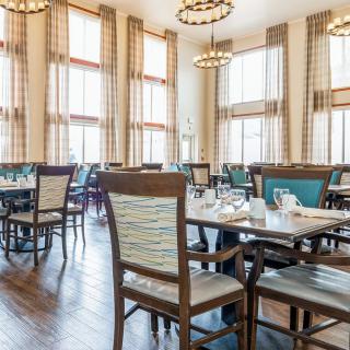 A spacious and well-lit dining room with neatly arranged tables and chairs, featuring large windows and elegant chandeliers.