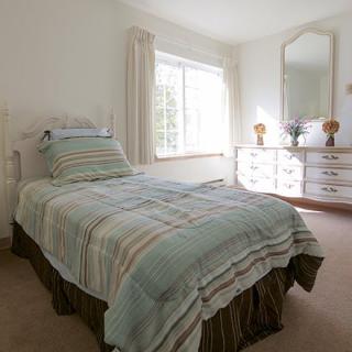 A cozy bedroom featuring a single bed with striped bedding, a dresser with a mirror, and large window with curtains.