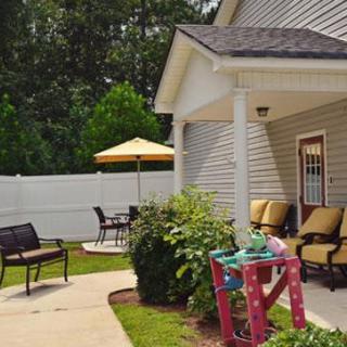 A cozy patio area with outdoor seating, including chairs and a table under an umbrella, surrounded by greenery and a white fence.