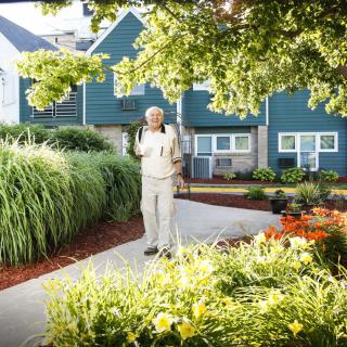An elderly man with a backpack stands on a garden pathway in front of a house surrounded by lush greenery and vibrant flowers on a sunny day.