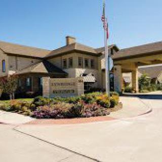 An inviting building entrance with a well-maintained driveway and lush landscaping under a clear, sunny sky.