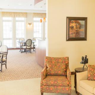 A bright and inviting seating area with comfortable chairs and artwork on the wall, leading to a dining space with multiple windows.