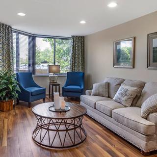 A cozy living room with a comfortable sofa, blue chairs, and a round coffee table, featuring hardwood floors and large windows.