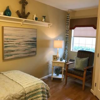 A cozy bedroom with nautical-themed decor, featuring a bed, a lamp on a nightstand, and a comfortable chair by the window.