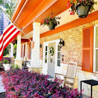 A charming porch with vibrant flowers and an American flag, featuring a rocking chair and hanging plants.