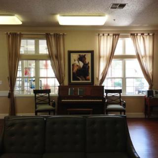 A cozy living room with a piano placed between two windows, adorned with curtains and an artwork above the piano.