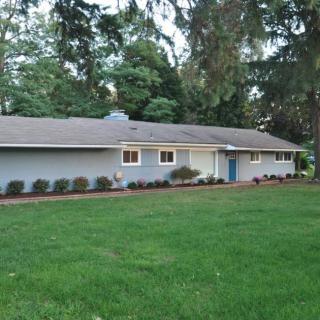 A single-story residential house with a large green lawn and surrounding trees.