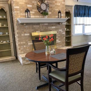 A cozy living room with a brick fireplace, a clock, and a round table with chairs and flowers.