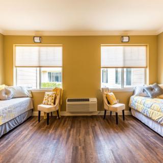 A bright bedroom featuring twin beds with yellow decor, wooden flooring, and two windows.