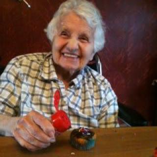 An elderly woman smiles while engaging in a painting activity indoors.