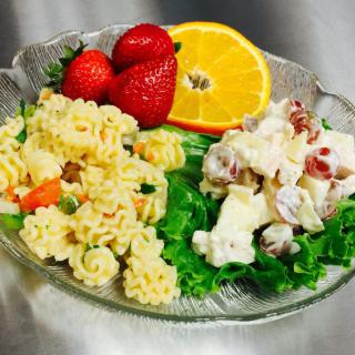 A plate featuring pasta salad with vegetables, potato salad on a bed of lettuce, strawberries, and an orange slice.