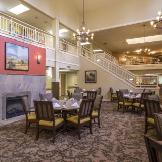 A spacious dining area with elegant chandeliers, neatly arranged tables and chairs, a carpeted floor, and a cozy fireplace.